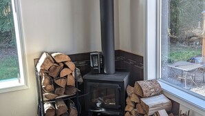 Une pièce chaleureuse avec un poêle à bois, une fenêtre donnant sur l’extérieur et une pile de bois de chauffage bien rangée.