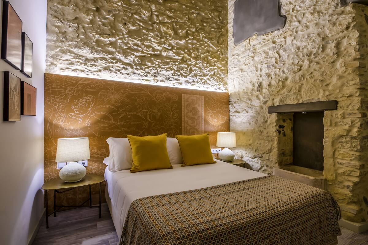 A bedroom with a stone wall, a bed with a patterned bedspread, two bedside lamps, and a fireplace.