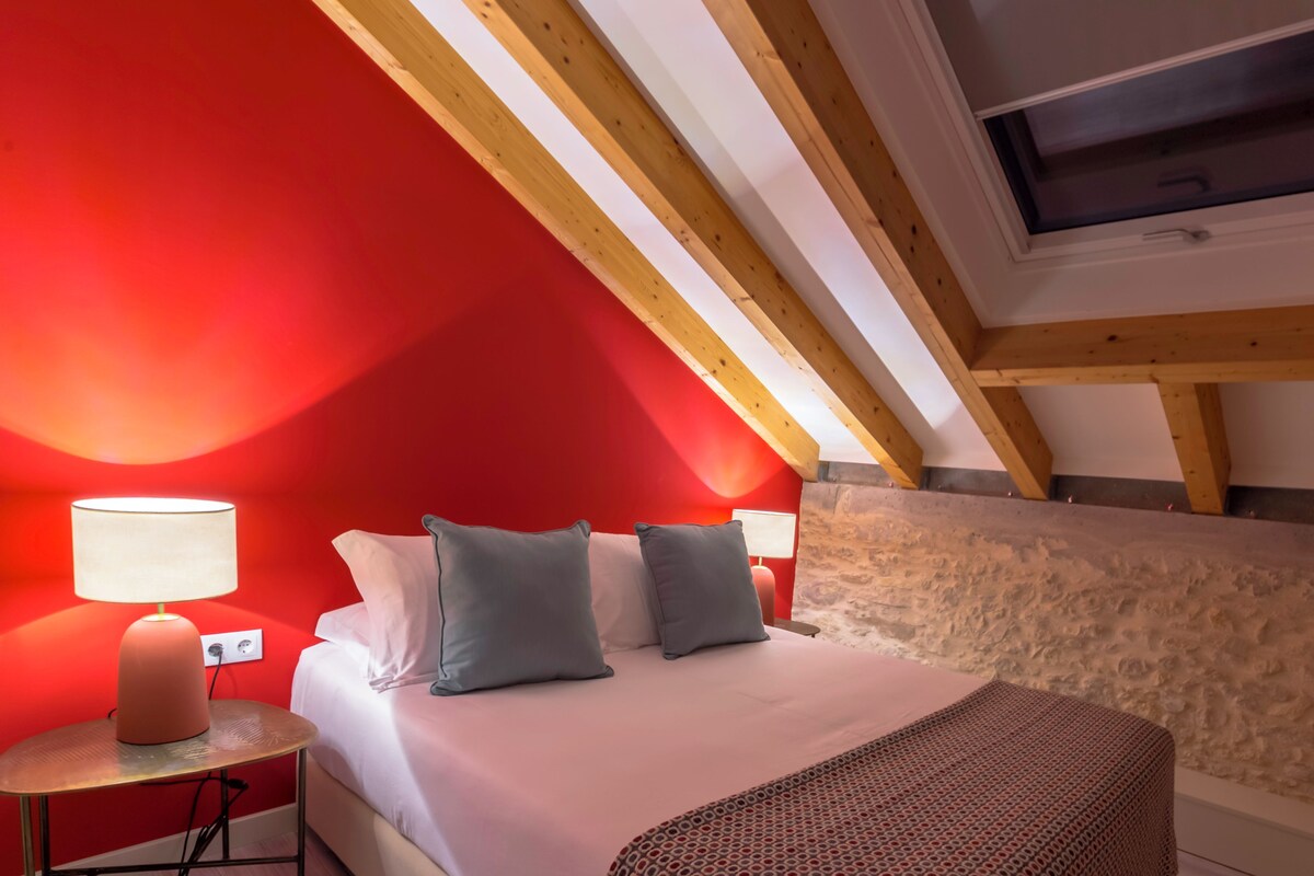 A bedroom with a sloped ceiling, a bed with pillows, a bedside table with a lamp, and a window.