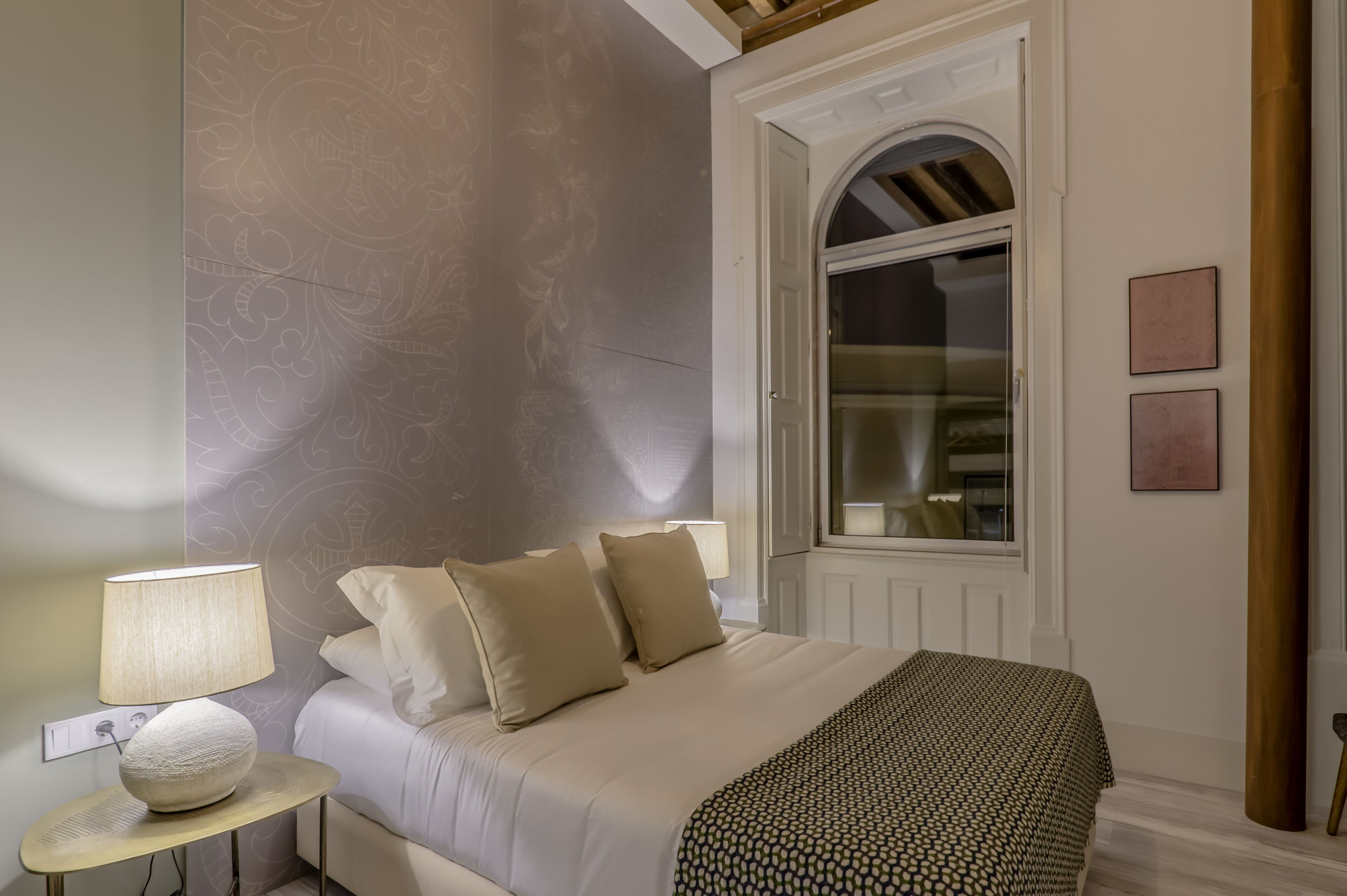 Un dormitorio con cama, lámparas de noche, una mesa redonda con lámpara y ventana con vistas.