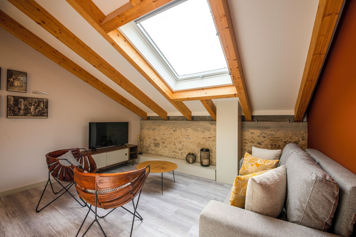 Uma sala de estar estilo loft com vigas de madeira aparentes, um claraboia, uma parede de pedra, uma televisão, um sofá e uma área de jantar com mesa e cadeiras.