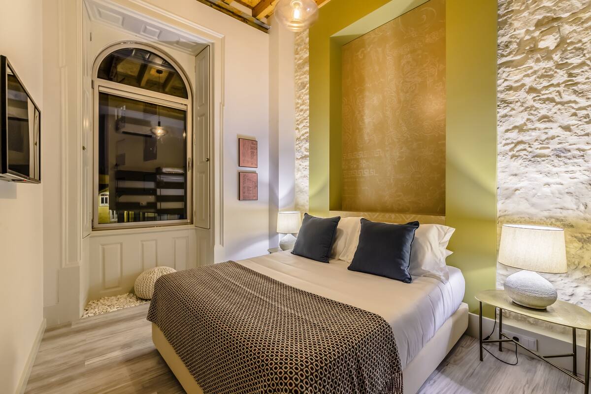 A bedroom with a bed, a bedside table with a lamp, a window, and a wall with a textured pattern.