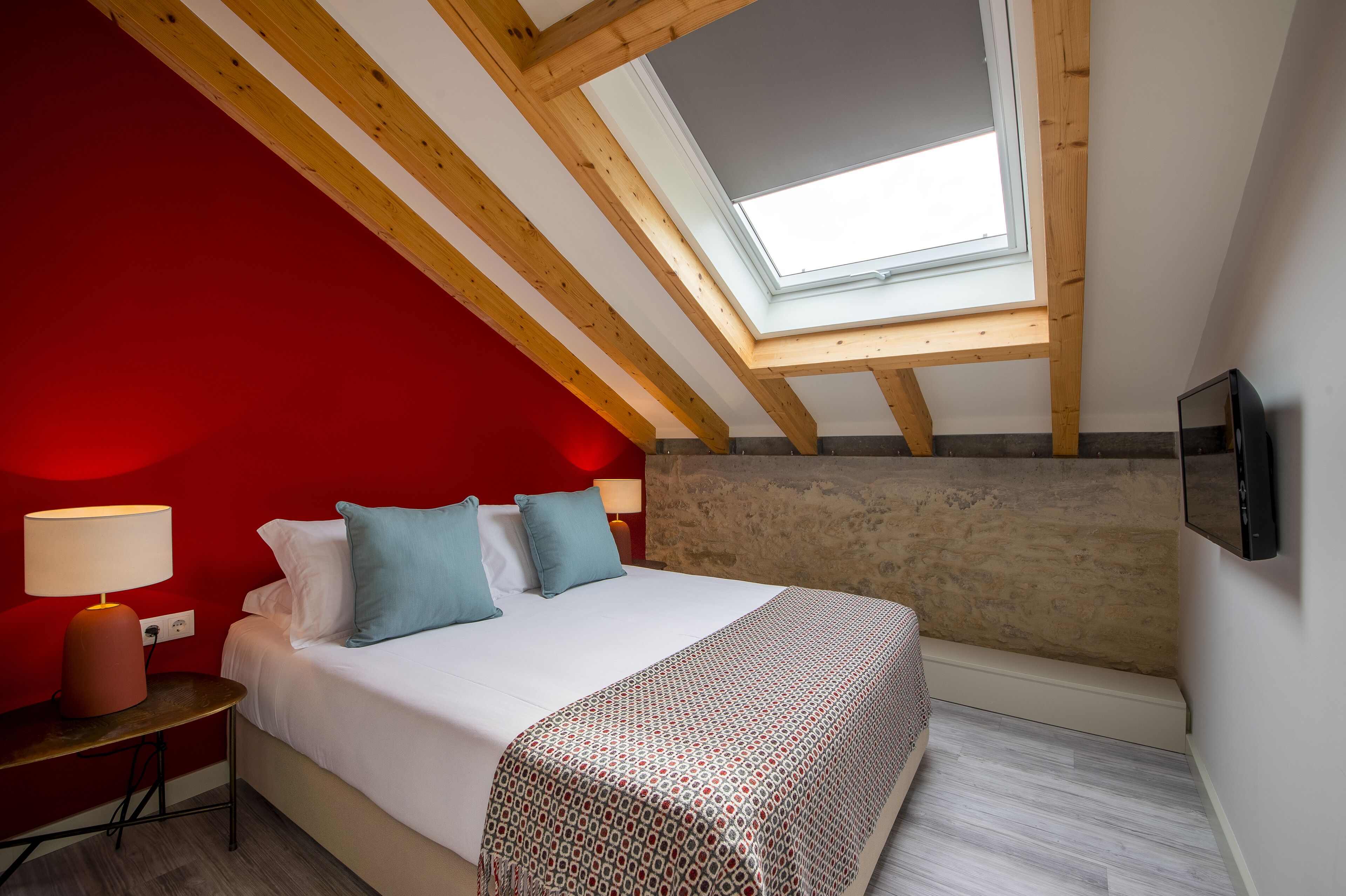 A bedroom with a sloped ceiling, a skylight, a bed with pillows, a bedside table with a lamp, and a patterned throw blanket.