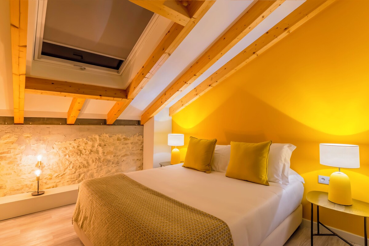 A bedroom with a sloped ceiling, a skylight, a bed with white bedding, two bedside lamps, and a stone wall.