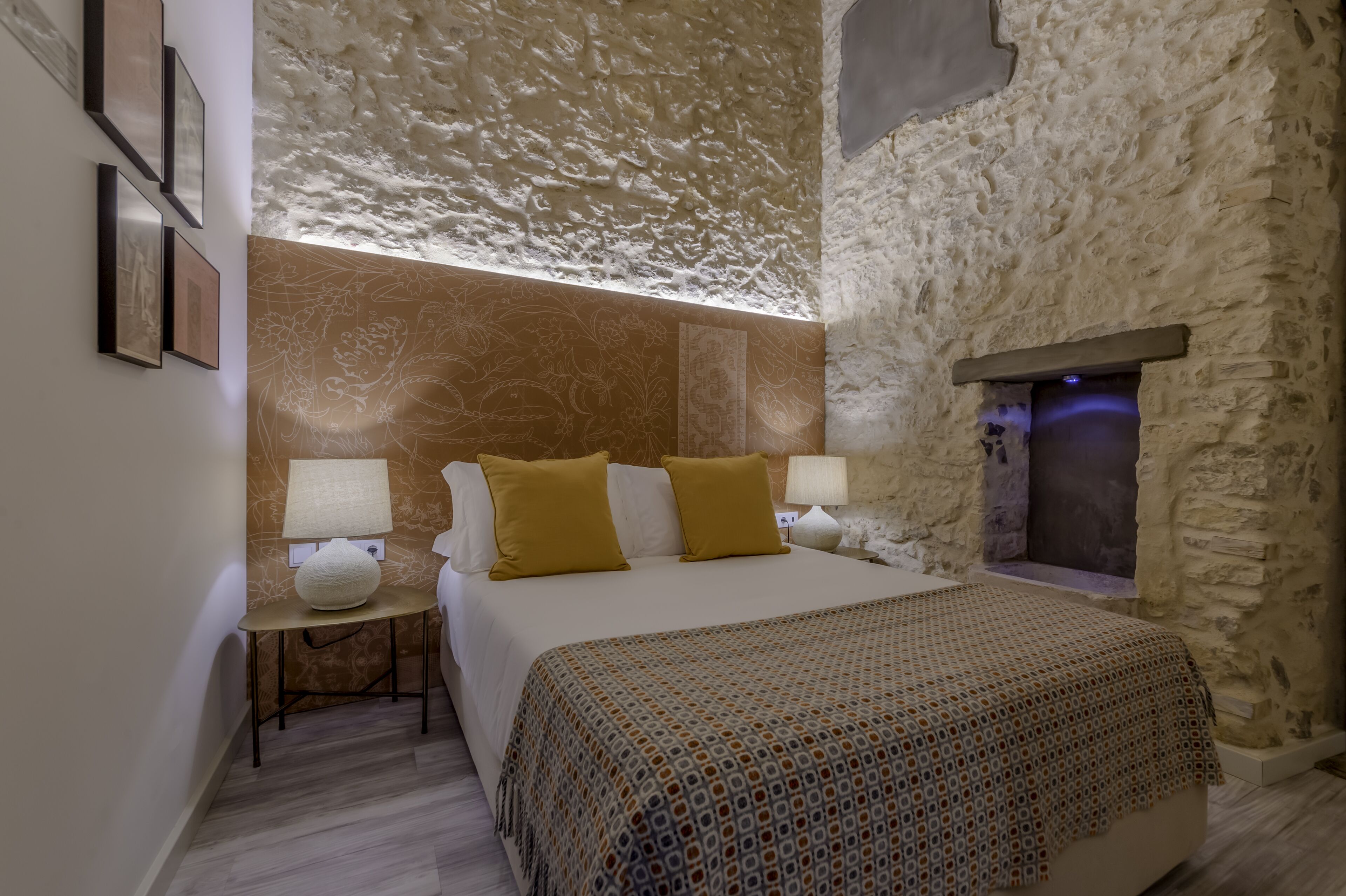A bedroom with a stone wall, a bed with two pillows, a bedside table with a lamp, and a patterned throw blanket.