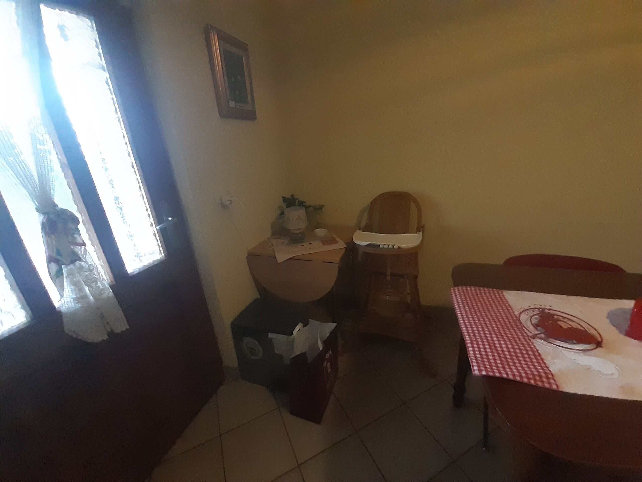 A small dining area with a table, chairs, and a high chair. There is a window with curtains, a door, and a trash bin.