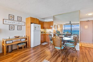 A compact kitchen with wooden cabinets, a white refrigerator, and a dining area with wicker chairs and a glass table.