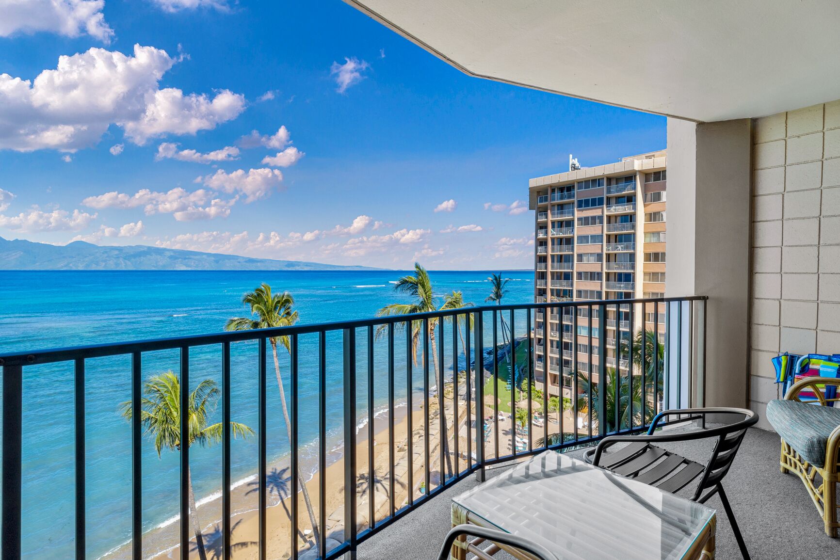 A balcony with a view of a beach and ocean, a table and chairs, and a high-rise building in the background.