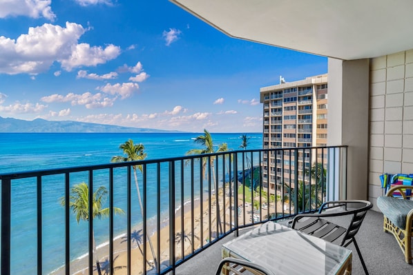 A balcony with a view of a beach and ocean, a table and chairs, and a high-rise building in the background.