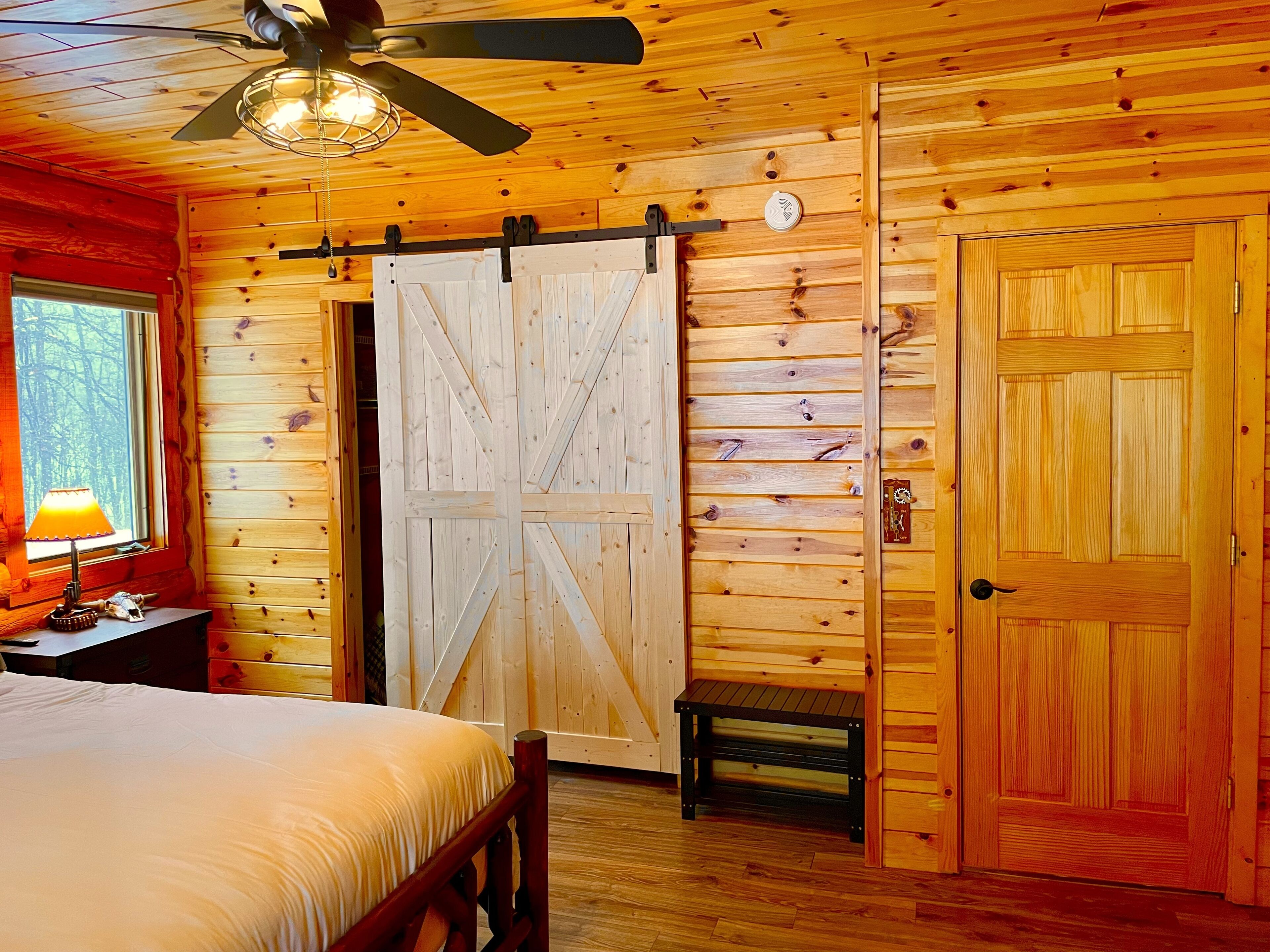 Ein Schlafzimmer in einer Holzhütte mit einem Bett, einem Nachttisch, einer Lampe, einem Deckenventilator und einer großen, strohgedeckten Tür.