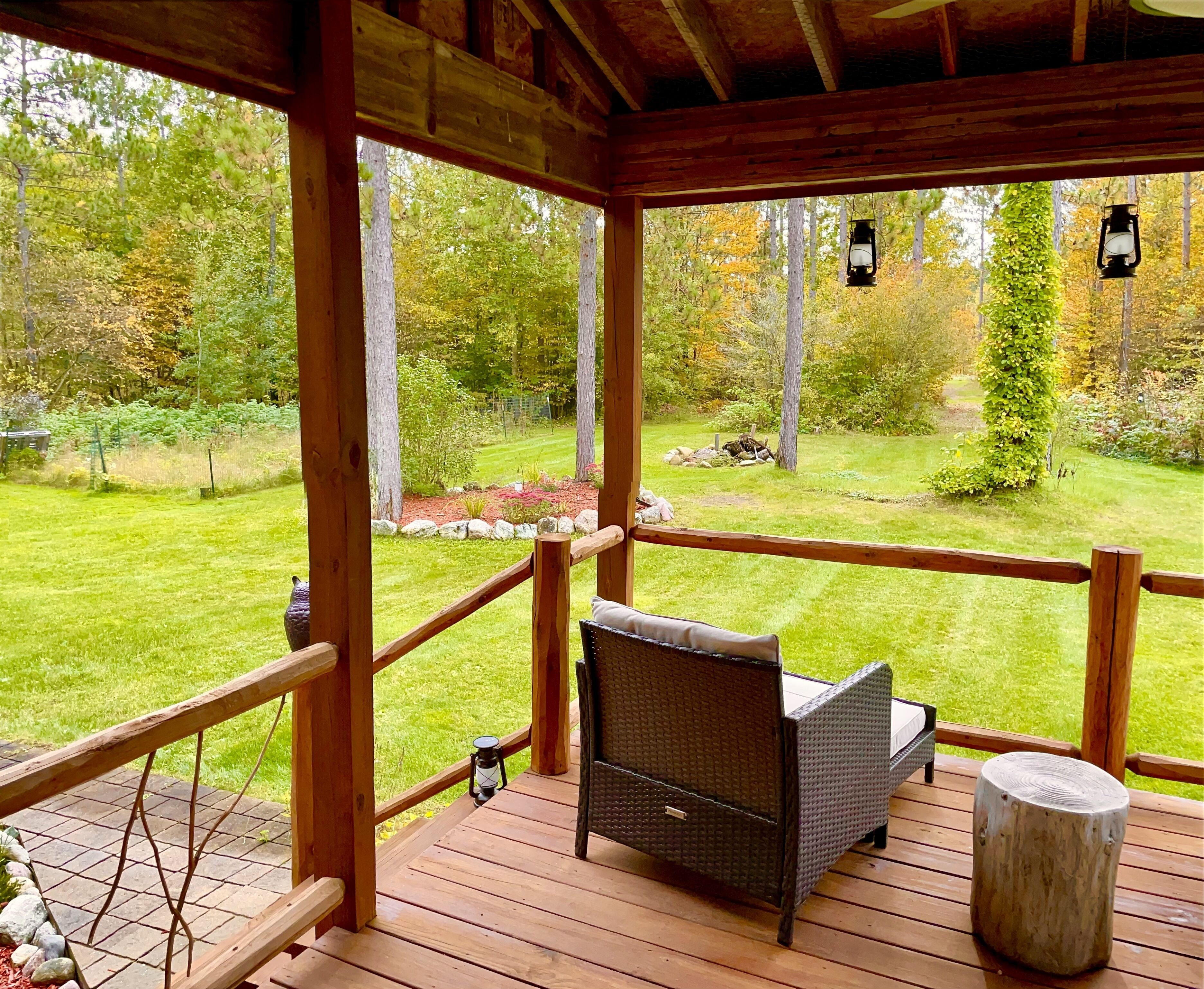 A covered outdoor deck with a wicker sofa and a stone table, overlooking a well-maintained lawn and forested area.