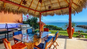 Un patio al aire libre con una mesa de vidrio, sillas azules y anaranjadas, una parrilla y vistas al océano.