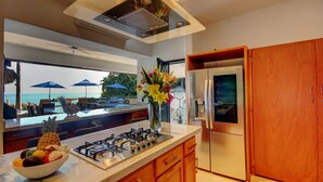Una cocina moderna con estufa a gas, refrigerador de acero inoxidable, armarios de madera y vista al océano a través de amplias ventanas.