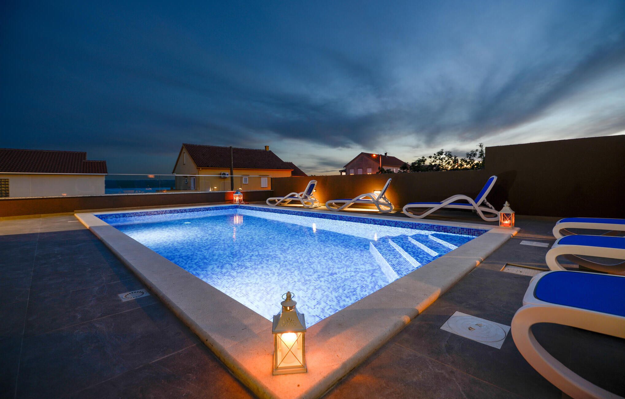 A rooftop swimming pool with lounge chairs and a lantern, surrounded by houses under a twilight sky.