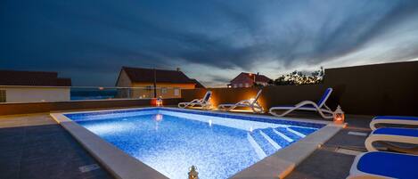 A rooftop swimming pool with lounge chairs and a lantern, surrounded by houses under a twilight sky.