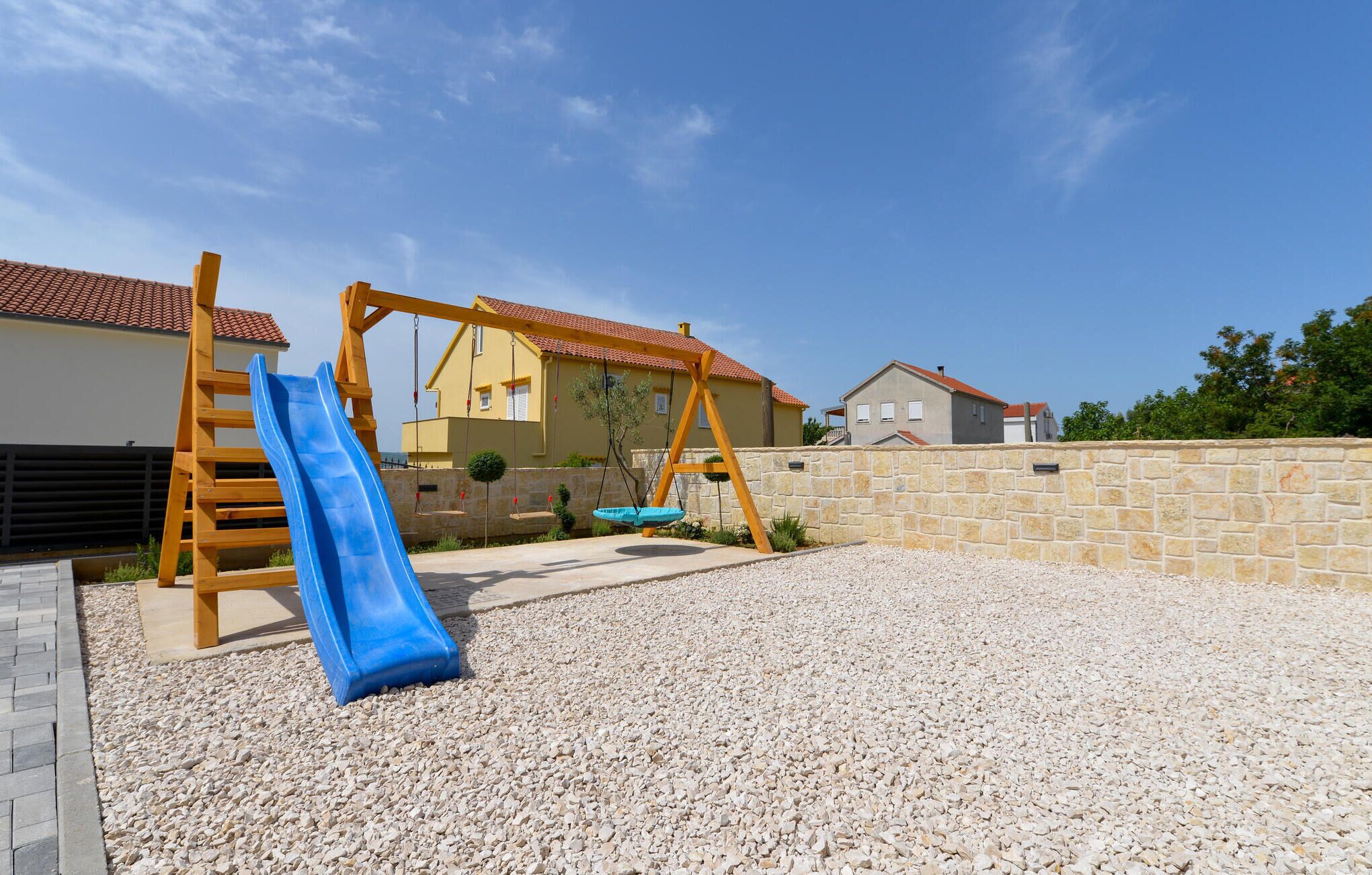 A playground with a blue slide and swings, surrounded by a stone wall and houses.