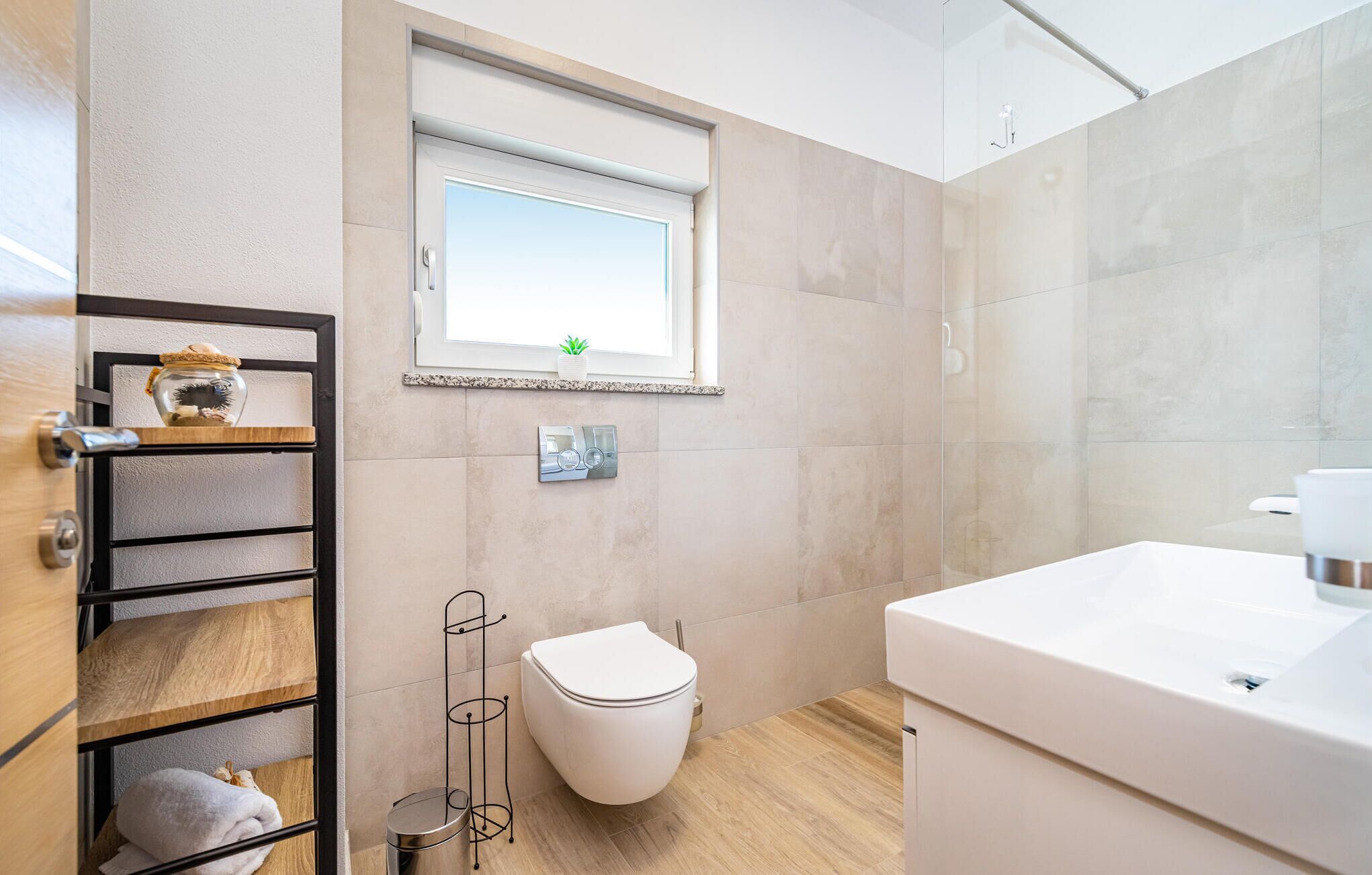 Un baño moderno con inodoro, lavamanos y una ventana sobre el inodoro. Hay una repisa con un tarro y una planta pequeña.