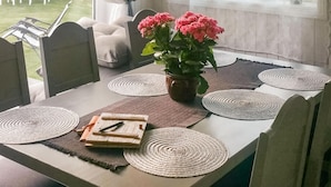 Un comedor con una mesa dispuesta para dos personas, con individuales tejidos, una planta en maceta con flores rosas y un libro y un bolígrafo sobre la mesa.