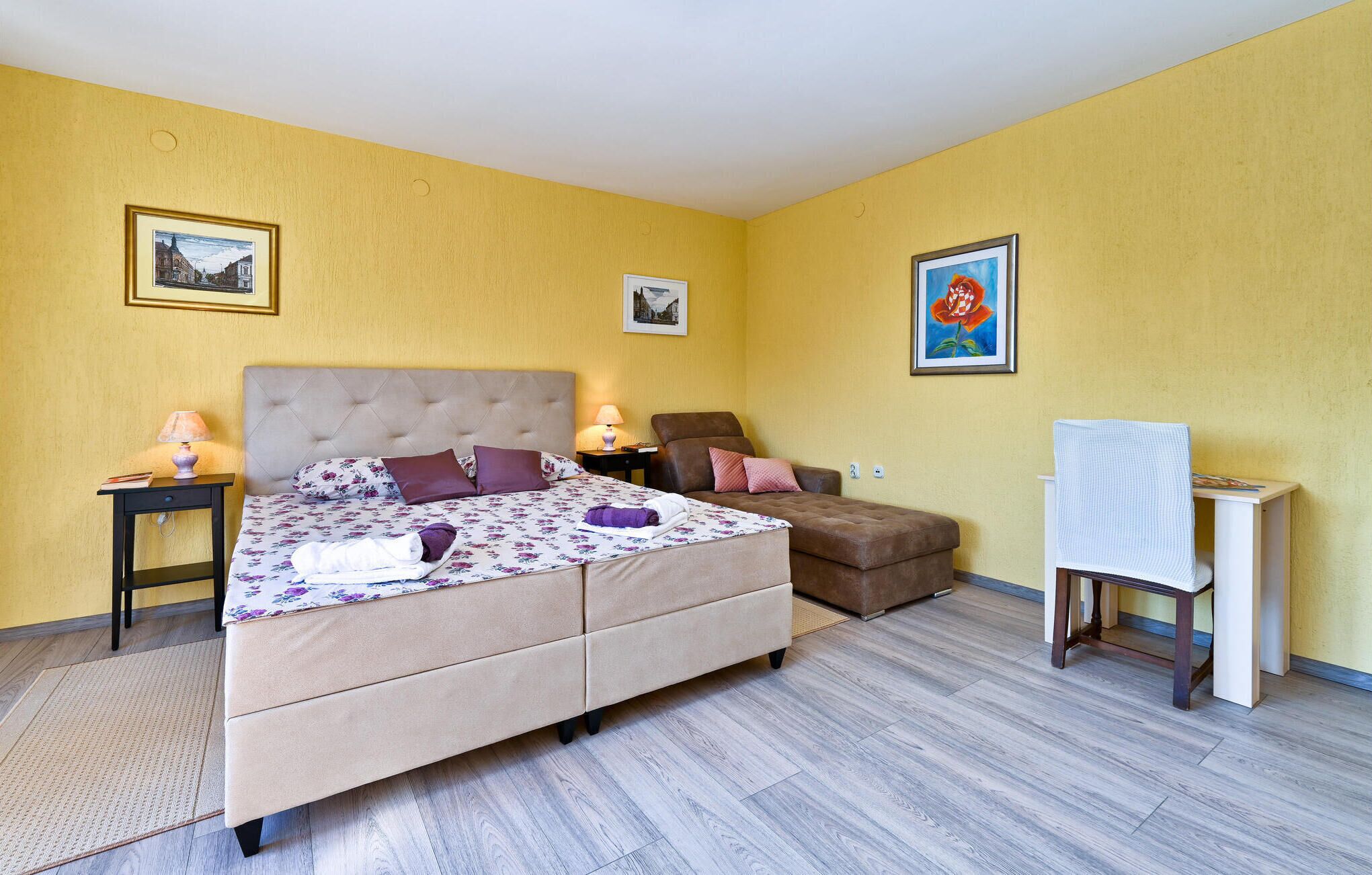 A bedroom with a bed, a chair, a desk, and two paintings on the walls.