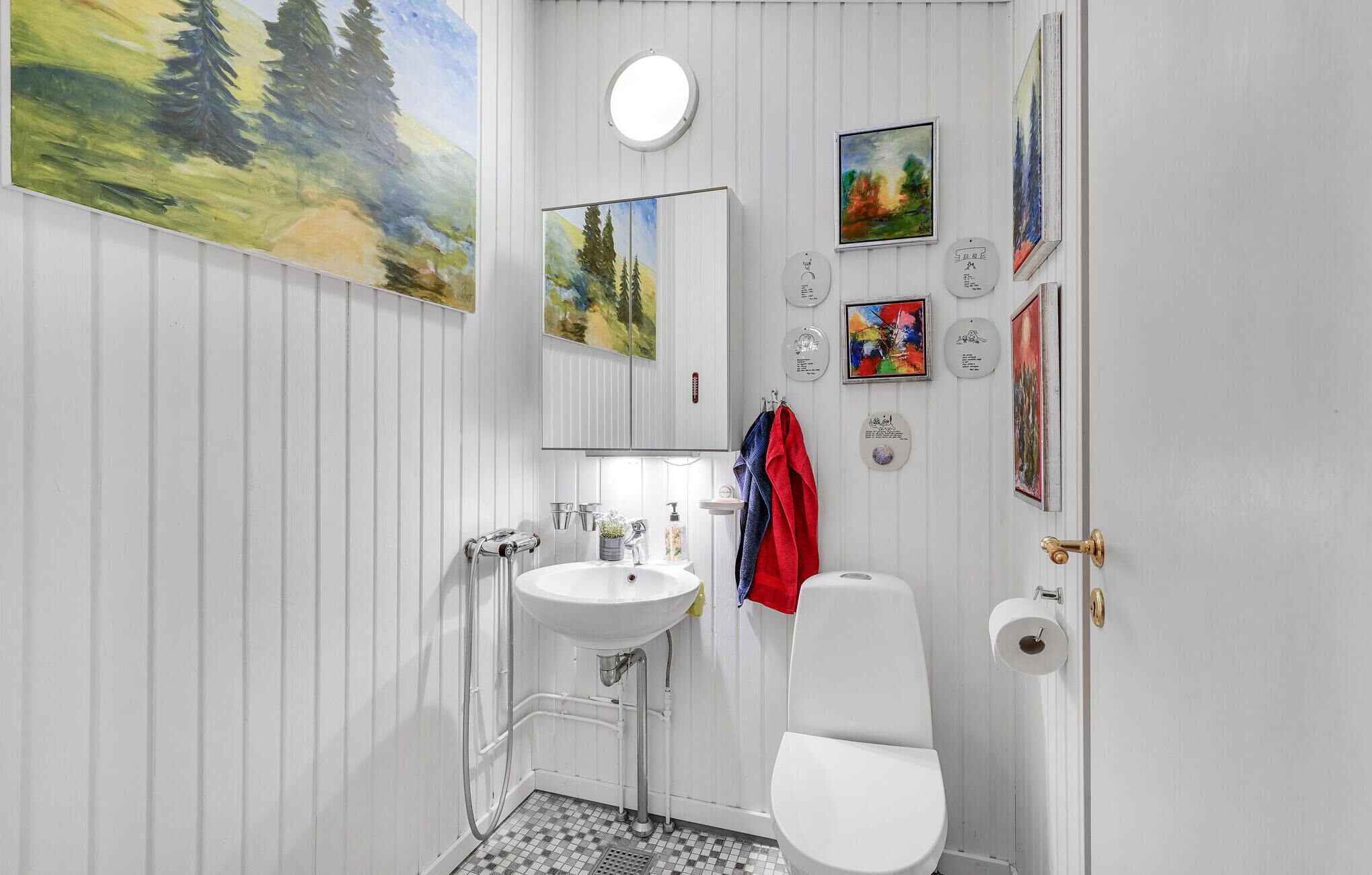Baño con lavamanos, inodoro y ducha blancos. Varias pinturas enmarcadas decoran las paredes.