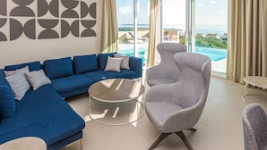 Un salón moderno con un sofá azul, un sillón estampado y una mesa de centro redonda. Amplias ventanas ofrecen vistas a una piscina y un paisaje pintoresco.