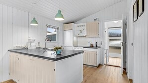 Et moderne køkken med hvide skabe, en central ø, hængende lamper og udsigt gennem en åben dør.
