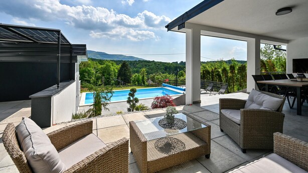 Un patio exterior moderno con mobiliario de mimbre, una piscina y vistas a un paisaje montañoso.