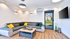 Una sala moderna con un sofá seccional gris, una mesa de centro de madera, luces empotradas en el techo y un ventanal con vistas a una casa y follaje.