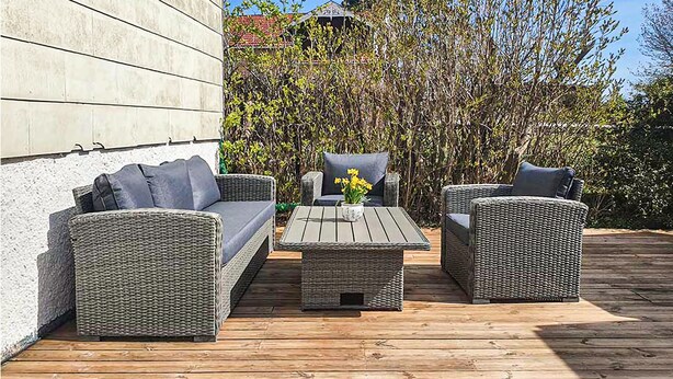 En udendørs terrasse med fletningsmøbler, heriblandt en sofa og to lænestole, et træbord og en lille potteplante.