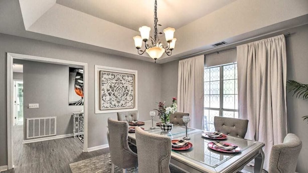 A dining room with a glass table, grey upholstered chairs, a chandelier, and a large window with curtains. - LUXURY GETAWAY, 4 BR, 2.5 BATH, NEAR FT. STEWART (Hinesville)