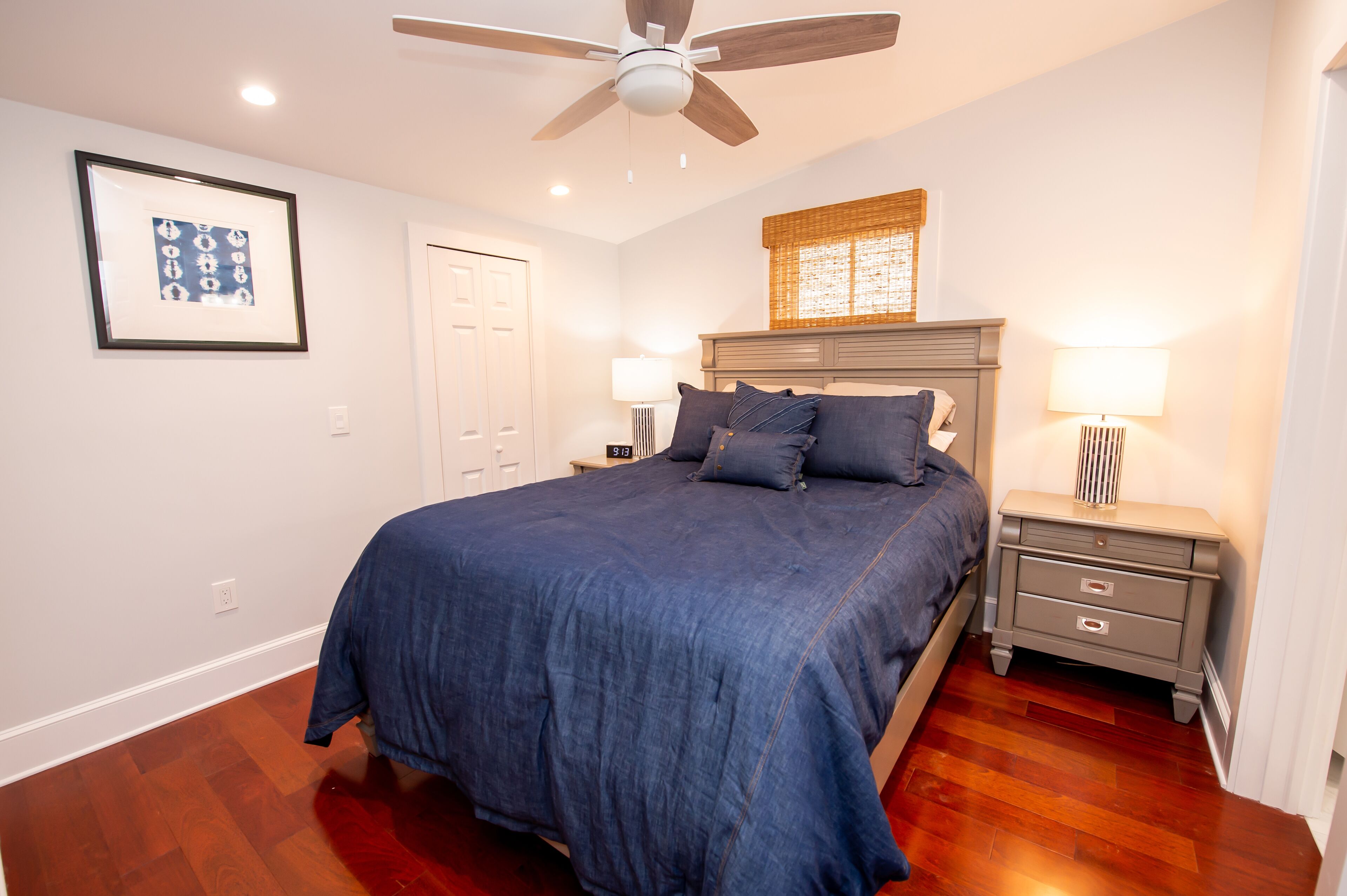 A bedroom with a bed, a nightstand, a ceiling fan, a wall-mounted lamp, and a framed picture.