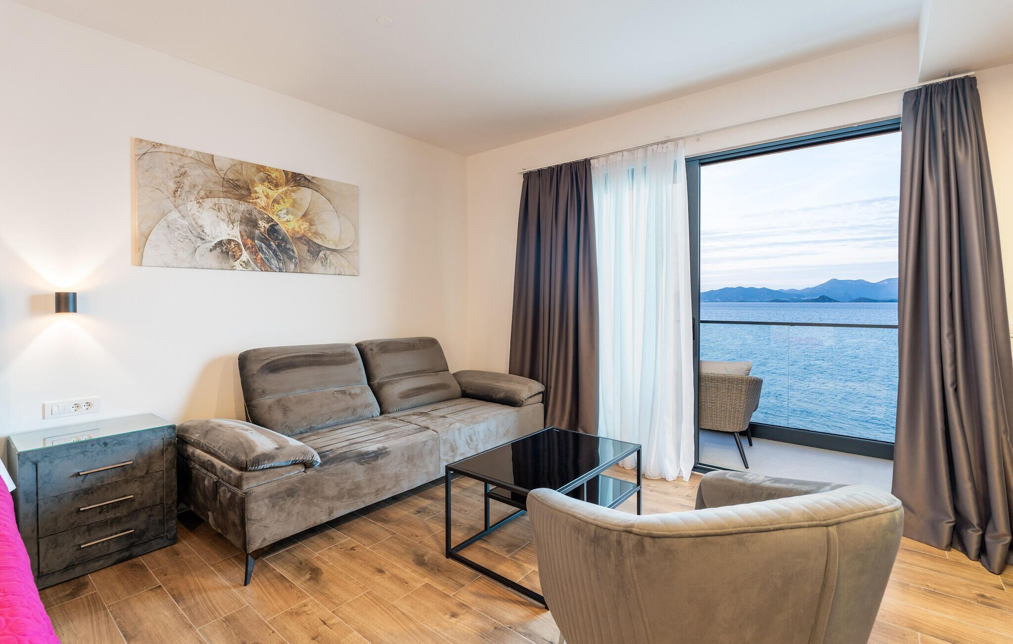 A modern living room with a sofa, a glass coffee table, a side table, and a view of the sea through large windows.