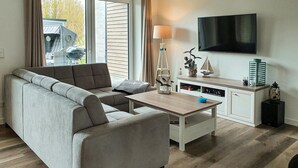 A modern living room with a grey sofa, a wooden coffee table, a TV mounted on the wall, and a kitchen area with cabinets and a stove. - Awesome home in Büsum with sauna (Büsum)