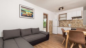 A modern living room with a grey sectional sofa, a dining area with a wooden table and brown chairs, a kitchen with white cabinets and a patterned backsplash, and a framed painting of boats on the wall. - Lovely apartment in Cres with WiFi (Cres)