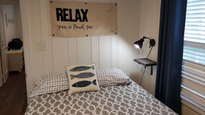 Un dormitorio con cama, almohadas, un letrero en la pared, un escritorio con lámpara y una ventana con cortinas.