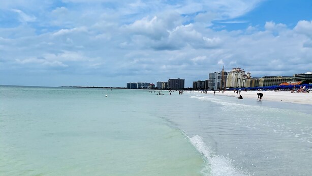 Ein Strand mit klarem, flachen Wasser, sandigem Ufer und Gebäuden im Hintergrund.