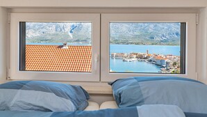 Une chambre avec deux fenêtres donnant sur une ville côtière et un paysage montagneux.