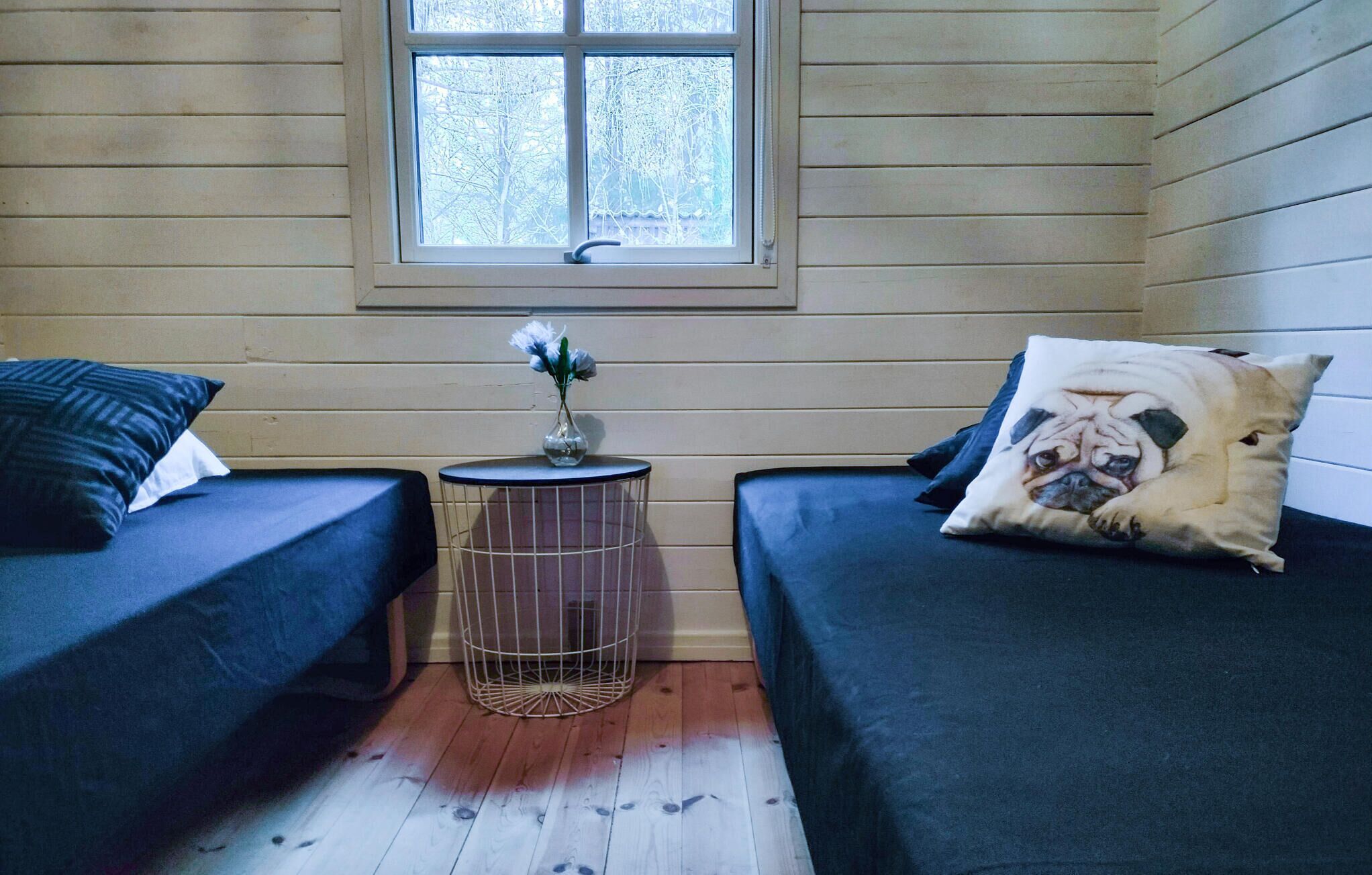 A room with two beds, a small round table with a vase of flowers, and a window with a view of trees.