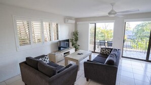 A modern living room with a sofa set, a coffee table, and a TV. There is a balcony with a view of greenery.