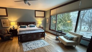 A bedroom with a large bed, a ceiling fan, a window with blinds, a bedside table with lamps, a chair, and a sofa. - Downtown Fish Creek Luxury Getaway 3BR/2.5BA, Game Room, Elevator & Deck Views! (Fish Creek)