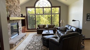 A living room with a stone fireplace, leather sofas, a coffee table, and a large window with a view of trees.