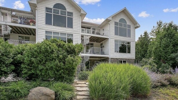 A two-story house with a balcony on the second floor, a stone pathway leading to the entrance, and a well-maintained garden with various shrubs and a large rock.