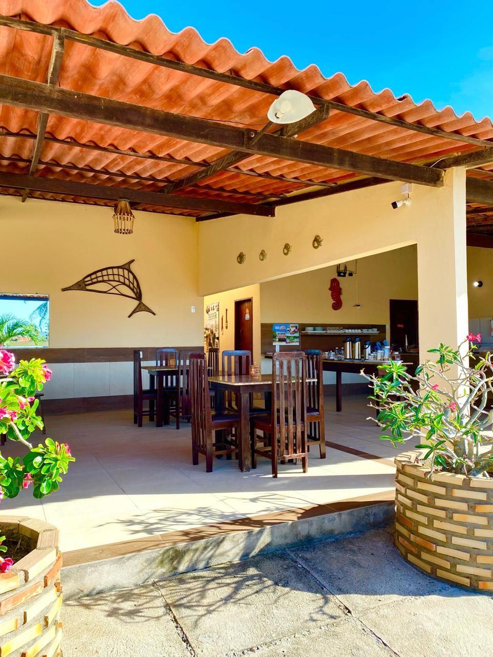 Uma área de pátio externa com cobertura de azulejos, mesas e cadeiras de madeira, arte decorativa de peixes na parede e vasos com plantas.
