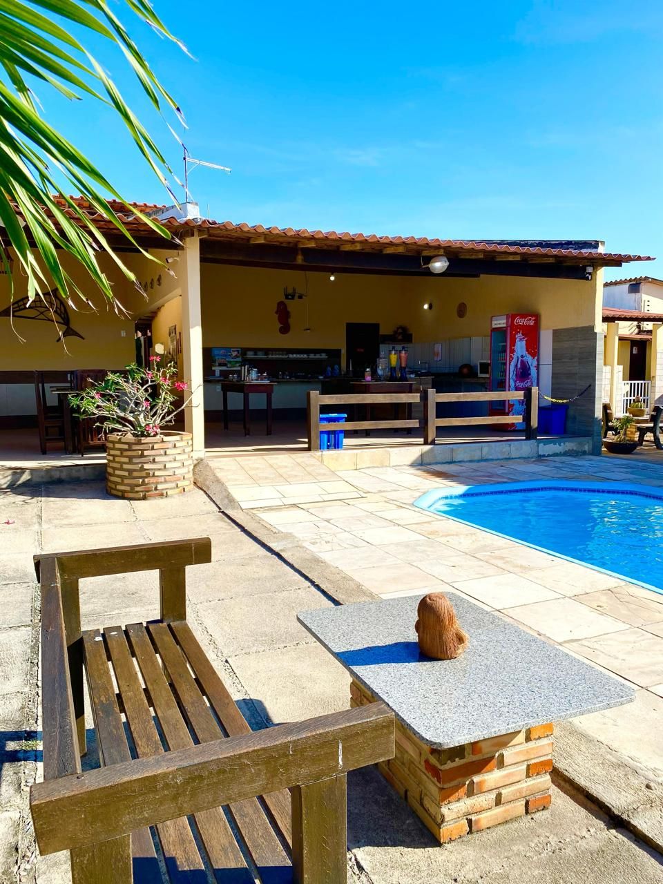 Zona de piscina con un banco de madera, una mesita y un refrigerador de Coca-Cola.