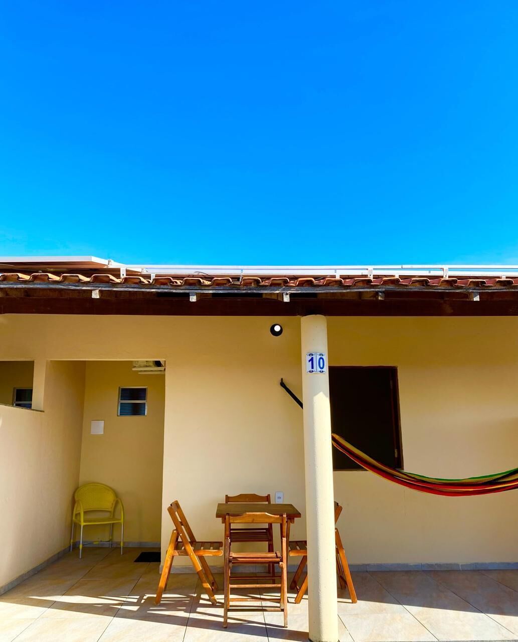 Un espacio al aire libre cubierto con suelo de baldosas, una mesa y sillas de madera, una silla amarilla y una hamaca.