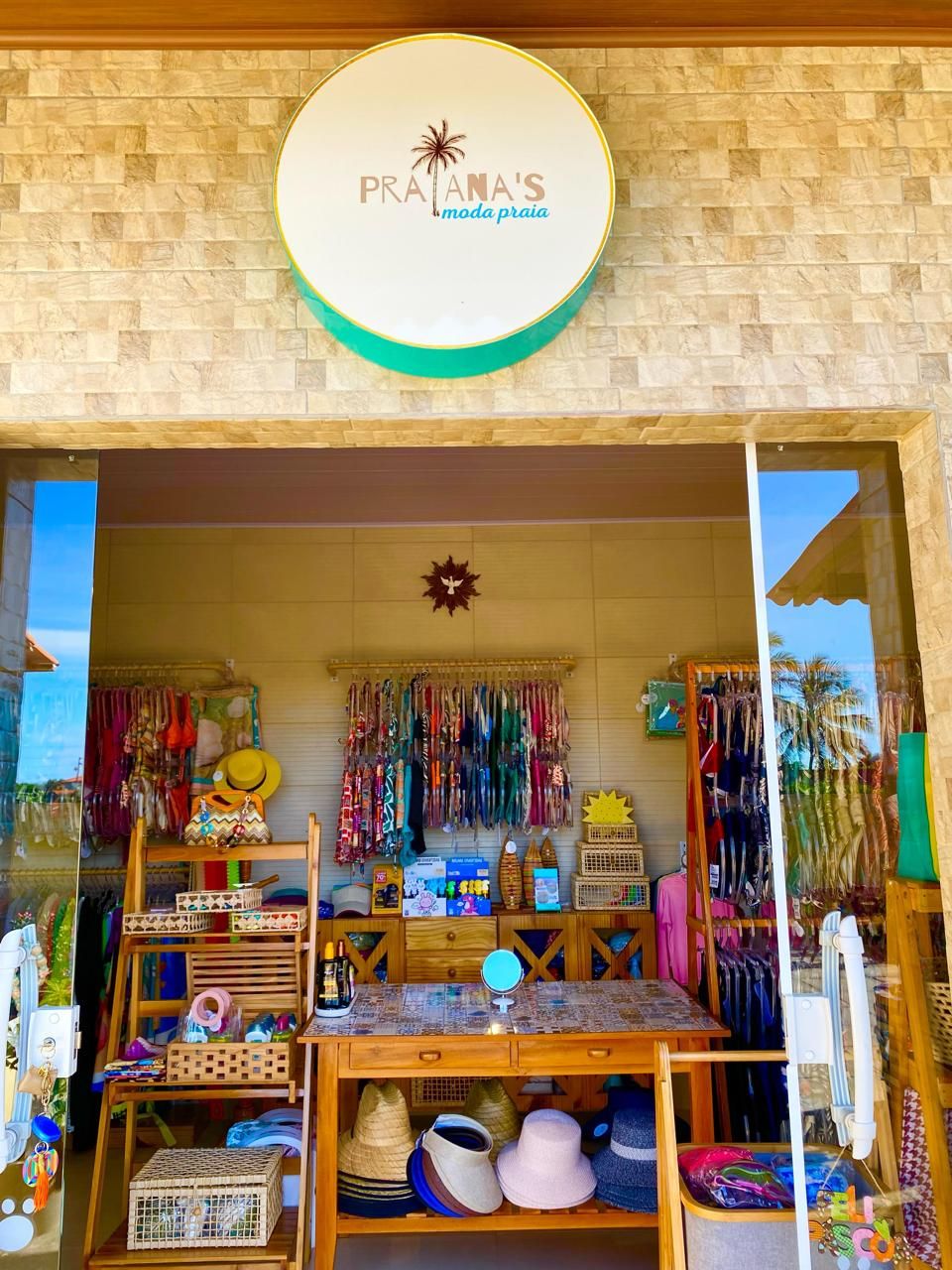 Una fachada con un letrero que dice "PRA'TANA'S moda praia" y varios accesorios de playa coloridos exhibidos afuera.