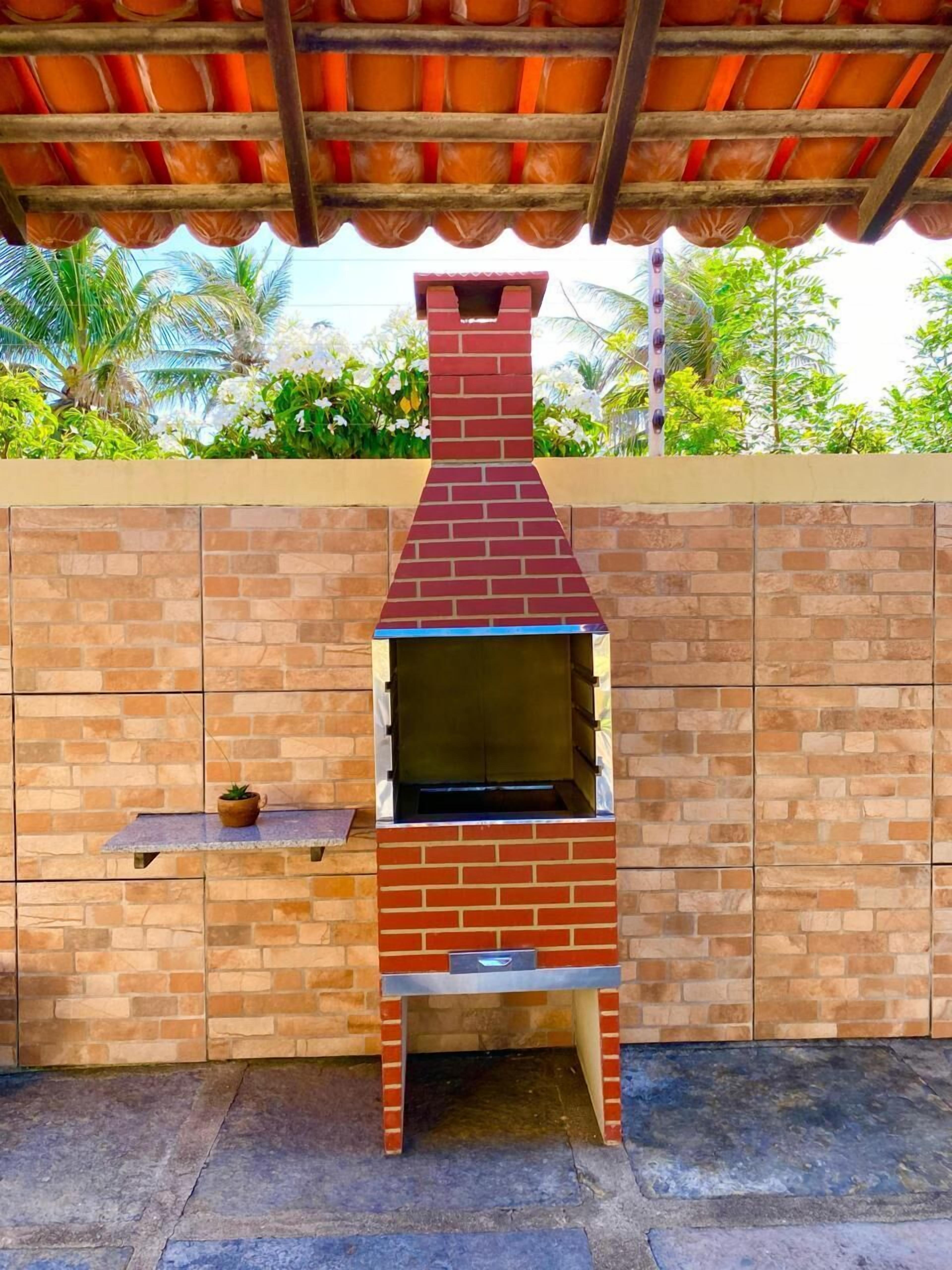 An outdoor brick oven with a chimney, set against a brick wall with a small shelf and potted plant.