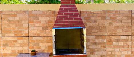 An outdoor brick oven with a chimney, set against a brick wall with a small shelf and potted plant.