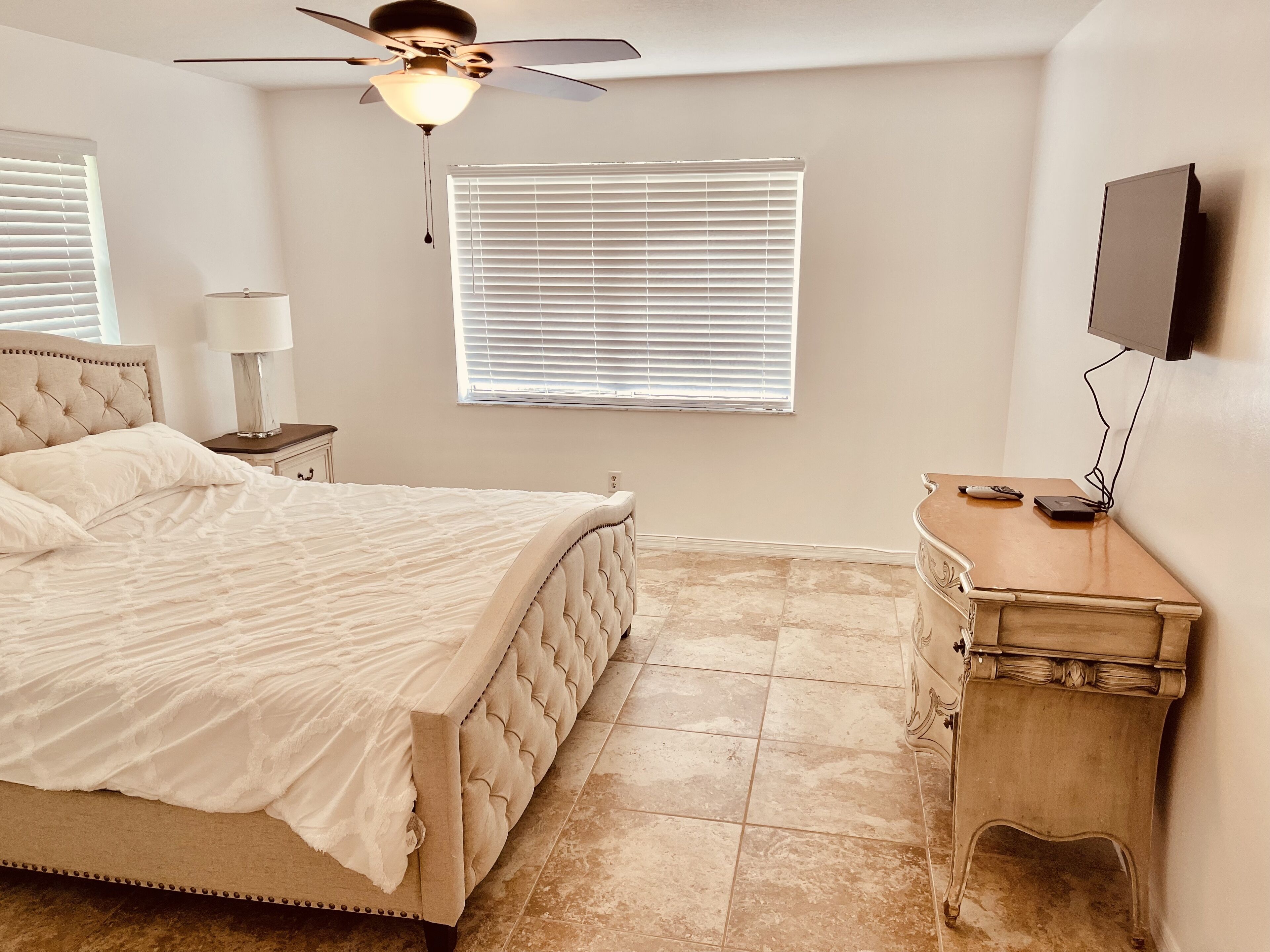 A bedroom with a bed, a nightstand, a dresser, a ceiling fan, a window with blinds, and a TV mounted on the wall.
