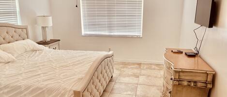 A bedroom with a bed, a nightstand, a dresser, a ceiling fan, a window with blinds, and a TV mounted on the wall.